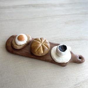 May include: Miniature breakfast set on a wooden serving board. The set includes a slice of toast with a fried egg, a round bread roll, and a small cup of coffee on a saucer. The items are painted in realistic colors.