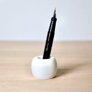 Op de afbeelding: Een witte keramische pennenhouder in de vorm van een appel met een zwarte vulpen erin.
