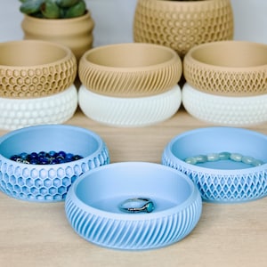May include: Four light blue decorative bowls with different textured patterns. Two bowls contain jewelry, one with a blue beaded bracelet and the other with a silver ring. The bowls are on a light wood surface.