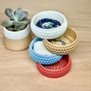 May include: Four stacked, round, decorative bowls with a honeycomb pattern. The bowls are in shades of white, beige, blue, and red. The top bowl has a blue beaded necklace and a ring inside.