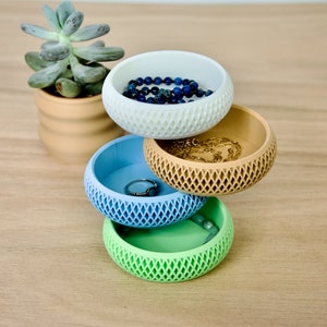 May include: Four stacked, round, decorative bowls with a geometric pattern. The bowls are in shades of white, beige, light blue, and green. The top bowl contains a blue beaded necklace, the second bowl contains a gold chain necklace, the third bowl contains a ring, and the bottom bowl contains a bracelet.