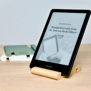 May include: A black Kindle e-reader with a white screen displaying the text "Handpicked reads from the Amazon Book Editors" and the Kindle logo. The e-reader is on a light brown stand.