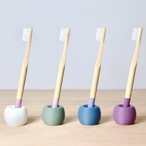 May include: Four bamboo toothbrushes with white bristles stand upright in four different coloured holders. The holders are white, green, blue, and purple.