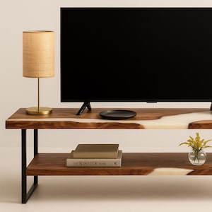 May include: A modern, two-tiered wooden TV stand with a live edge design and a white resin inlay. The stand features a black TV, a lamp, a black plate, books, and vases with yellow flowers. The stand is supported by black metal legs.