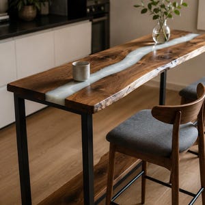 May include: A live edge wooden bar table with a white resin river design. The table has black metal legs and is paired with two wooden bar stools with gray upholstered seats. A cup and vase of greenery are on the table.