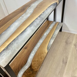 May include: A two-tiered console table with a unique design. The table features a wooden surface with a river of white and gray resin. The frame is black metal. The table is approximately 48 inches long, 12 inches wide, and 30 inches high.