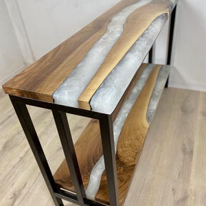 May include: A handcrafted console table with a live edge wood top and a river of white epoxy resin. The table has a black metal frame and a lower shelf. The wood has a natural grain pattern.