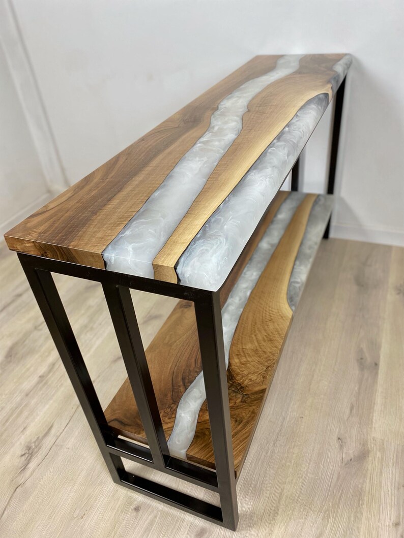 May include: A handcrafted console table with a live edge wood top and a river of white epoxy resin. The table has a black metal frame and a lower shelf. The wood has a natural grain pattern.