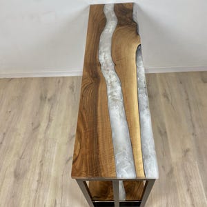 May include: A wooden bench with a live edge design and a river of white resin running through the center. The bench has a dark metal frame. The wood has a natural grain and a warm brown color.