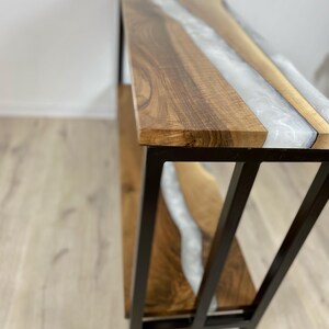 May include: A wooden console table with a live edge design and a river of white epoxy resin. The table has a black metal frame. The wood is a warm brown color.