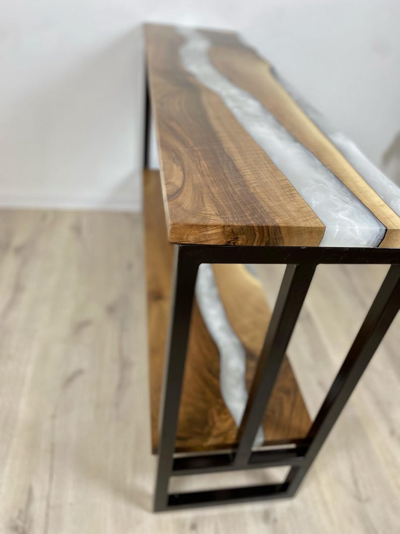 May include: A wooden console table with a live edge design and a river of white epoxy resin. The table has a black metal frame. The wood is a warm brown color.