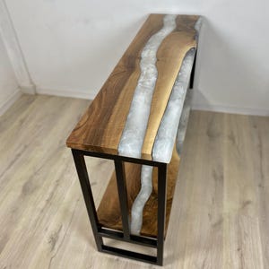 May include: A wooden console table with a live edge design and a river of white resin. The table has a dark wood finish and a black metal frame. The table is approximately 48 inches long.