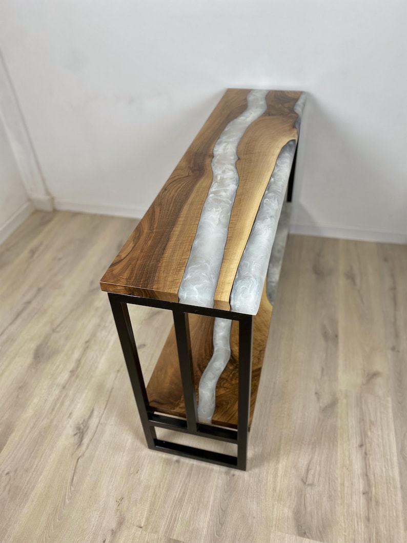 May include: A wooden console table with a live edge design and a river of white resin. The table has a dark wood finish and a black metal frame. The table is approximately 48 inches long.