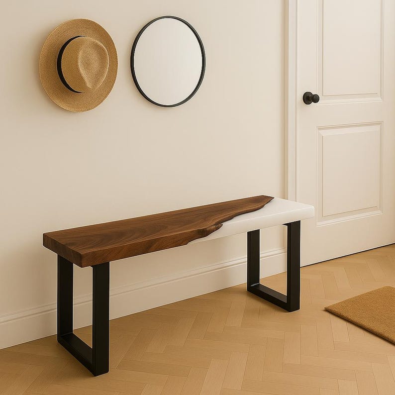 May include: A modern bench with a live edge walnut wood top and a white resin section. The bench has black metal legs. A straw hat and a round black framed mirror hang on the wall above the bench.