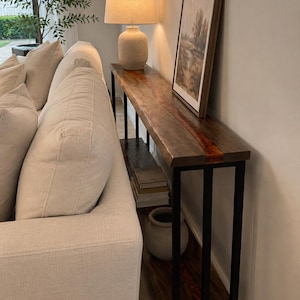 Puede incluir: Una consola de madera con estructura de metal negro, coronada por una lámpara y un cuadro de paisaje enmarcado. La mesa tiene un acabado de vetas de madera natural con un acento marrón rojizo. Un sofá con cojines color crema está a la izquierda.