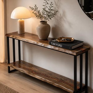 Könnte beinhalten: Eine Holzkonsolentisch mit einer Tischplatte mit rohem Rand und einem unteren Regal, das von schwarzen Metallbeinen getragen wird. Eine beige Lampe mit rundem Schirm und eine strukturierte Vase mit Grün stehen auf dem Tisch. Ein Buch mit dem Text "TOM FORD" liegt ebenfalls auf dem Tisch.