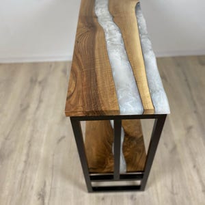 May include: A handcrafted console table with a live edge walnut top and a white epoxy resin river. The table has a black metal base and a natural wood grain finish. The table is approximately 48 inches long.