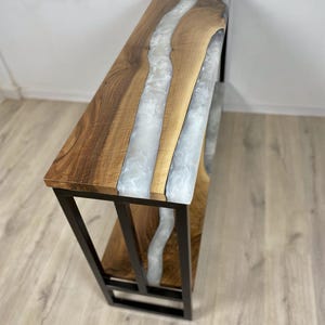May include: A wooden console table with a river of white resin flowing through the center. The table has a dark metal frame and a lower shelf. The wood has a natural grain, and the resin has a glossy finish.
