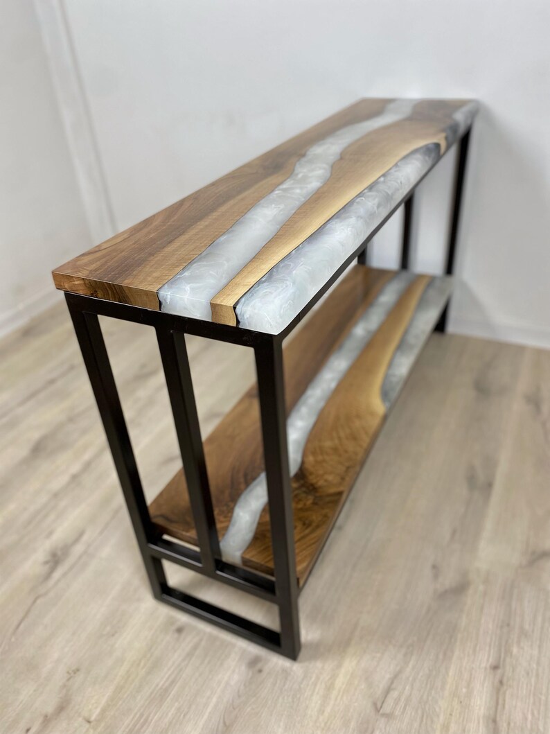 A modern console table with a black metal frame and two wooden shelves. The shelves feature a unique design with a river of white resin flowing through the center of the wood. The wood has a natural grain pattern, and the resin adds a contrasting element. The table is a stylish piece of furniture.