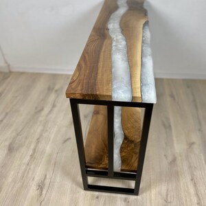 May include: A wooden console table with a live edge design and a river of white epoxy resin. The table has a black metal frame and a lower shelf. The wood has a natural grain.
