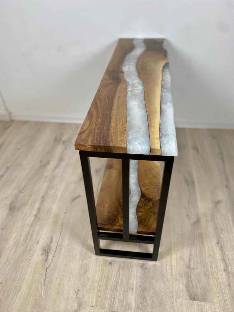 May include: A wooden console table with a live edge design and a river of white epoxy resin. The table has a black metal frame and a lower shelf. The wood has a natural grain.