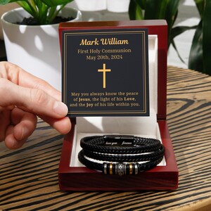 Könnte beinhalten: Eine schwarze Geschenkbox mit einem goldenen Kreuz auf dem Deckel. Die Box enthält ein schwarzes Lederarmband mit einem goldenen Verschluss und eine kleine Karte mit der Aufschrift "Mark William Erste Heilige Kommunion 20. Mai 2024" und einer Botschaft über den Frieden Jesu.