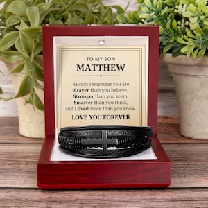 May include: A black leather bracelet with a silver cross is displayed inside a red wooden box. The box contains a card with the text "TO MY SON MATTHEW" and a loving message. The bracelet is a thoughtful gift.