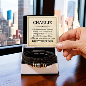 Könnte beinhalten: Ein schwarzes Lederarmband mit silbernen Akzenten wird in einer weißen Box mit einer personalisierten Nachricht geliefert, die lautet: "CHARLIE Denk immer daran, dass du mutiger bist, als du glaubst, stärker, als du scheinst, klüger, als du denkst, und geliebter, als du weißt. ICH LIEBE DICH FÜR IMMER".