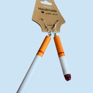 May include: Cigarette-shaped earrings with orange and white details, a red tip, and silver hooks. The earrings are displayed on a brown tag that reads "Handmade with love."
