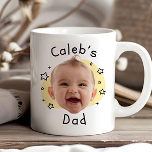 May include: White ceramic mug with a yellow circle and stars. The circle has a photo of a baby with the text "Caleb's Dad" below the photo.