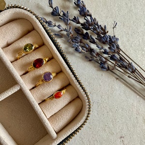 Peut inclure: Quatre bagues en or avec des pierres précieuses de différentes couleurs sont présentées dans un écrin à bagues en velours beige. Les bagues présentent une pierre précieuse verte, rouge, violette et orange.
