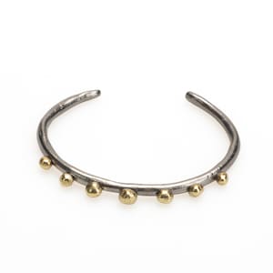 May include: A silver bangle bracelet with a hammered finish and gold accents. The bracelet has a simple, minimalist design with a row of small gold beads.