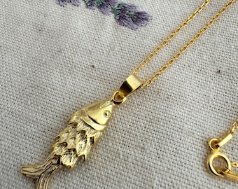 Puede incluir: Un collar de cadena dorada con un colgante de pescado detallado. La cadena es delicada y el pescado es un diseño realista.