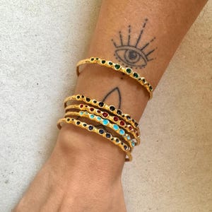 May include: A set of five gold bangles with colourful enamel accents. Each bangle features a row of small, round, enamel-filled circles in a different colour, including turquoise, red, black, and green.