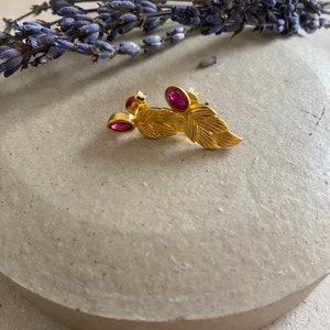 May include: Gold leaf earrings with two pink gemstones. The earrings are shaped like leaves and have a textured surface.