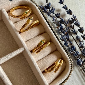 May include: Gold-toned snake rings with emerald and ruby accents are displayed in a jewelry case. The rings have textured scales and wrap around the finger. A sprig of lavender is in the background.