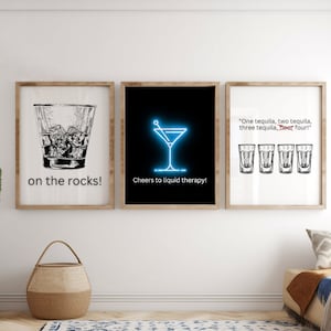 May include: Three framed prints with black and white illustrations of cocktails. The first print shows a glass of whiskey on the rocks with the text "on the rocks!" The second print shows a martini glass with the text "Cheers to liquid therapy!" The third print shows four shot glasses with the text "One tequila, two tequila, three tequila, floor four!"