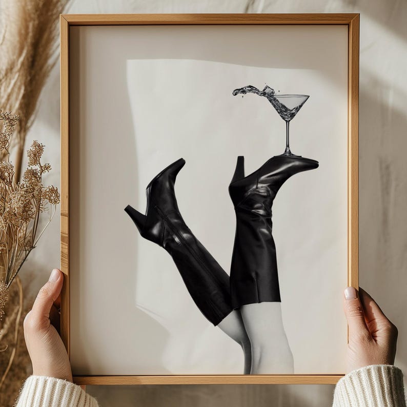 K&ouml;nnte beinhalten: Schwarzwei&szlig;foto von einem Paar schwarzer Stiefel mit einem Martini-Glas, das darauf balanciert ist. Die Stiefel sind angewinkelt und das Glas ist geneigt, was eine dynamische Komposition erzeugt.