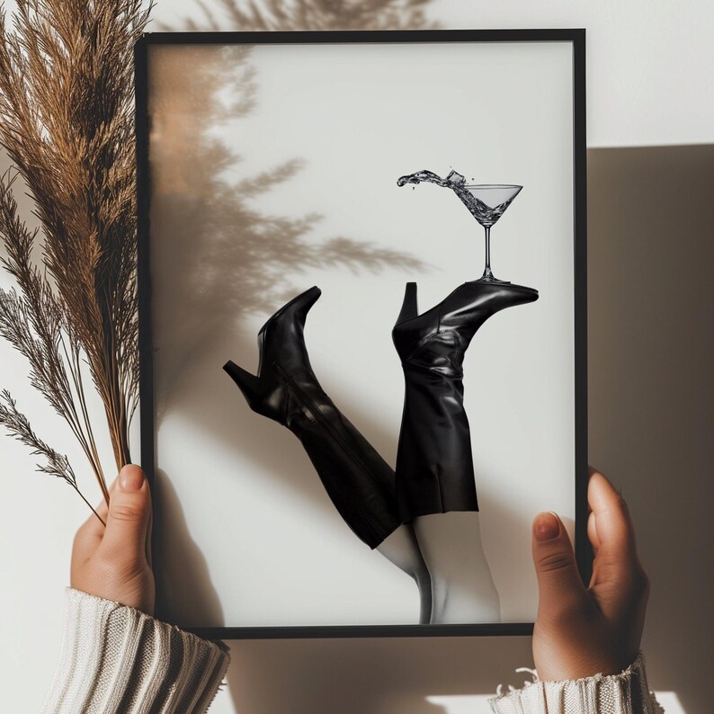 K&ouml;nnte beinhalten: Ein Schwarzwei&szlig;foto von einem Paar schwarzer Stiefel mit Abs&auml;tzen, die auf einem Martini-Glas stehen. Das Glas ist mit einer klaren Fl&uuml;ssigkeit gef&uuml;llt und hat einen Spritzer Fl&uuml;ssigkeit, der herausspritzt. Das Foto ist in einem schwarzen Rahmen gerahmt.