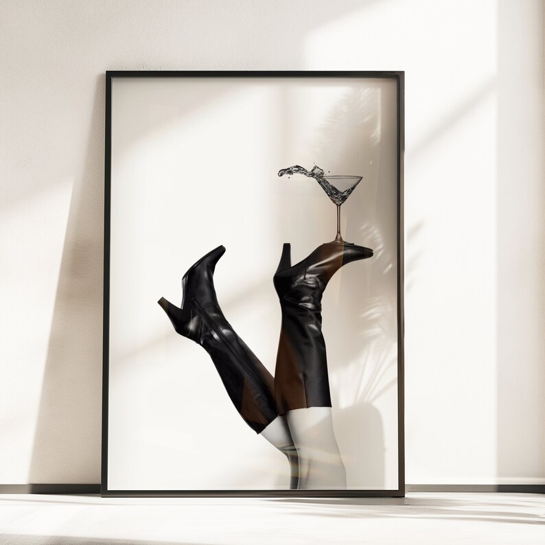 K&ouml;nnte beinhalten: Ein Schwarzwei&szlig;foto von einem Paar schwarzer Stiefel mit Abs&auml;tzen, wobei ein Stiefel ein Martini-Glas mit einem Spritzer Fl&uuml;ssigkeit h&auml;lt. Der Hintergrund ist wei&szlig;.