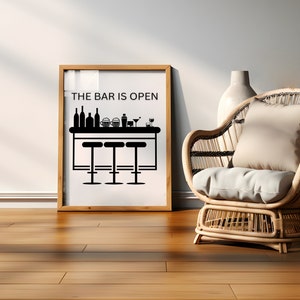 May include: A framed black and white illustration of a bar with stools and the text "THE BAR IS OPEN". The frame is wooden and has a white mat. The frame is sitting on a wooden floor. A wicker chair with a white cushion is in the foreground.
