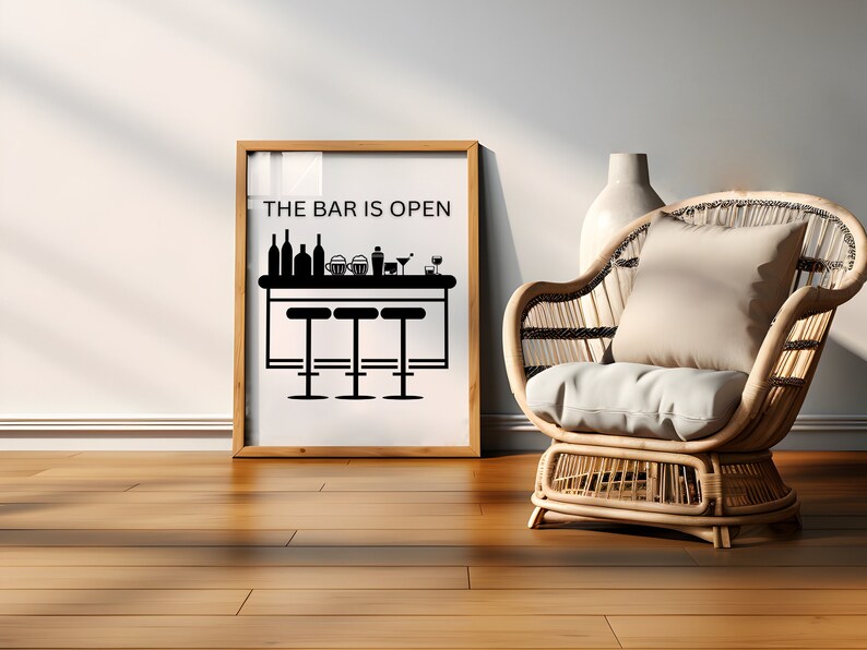 May include: A framed black and white illustration of a bar with stools and the text "THE BAR IS OPEN". The frame is wooden and has a white mat. The frame is sitting on a wooden floor. A wicker chair with a white cushion is in the foreground.