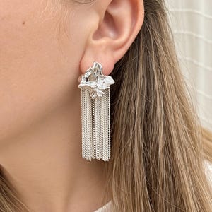 May include: A silver-toned earring featuring an irregular, molten-textured stud top with a cluster of multiple fine, dangling chain fringes that hang vertically from the base, creating a layered metallic effect against the earlobe.