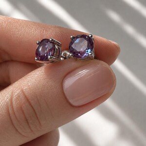 May include: A pair of stud earrings featuring round, faceted gemstones with a deep purple hue. The earrings are set in a silver metal, and are held by a hand with a neutral-toned manicure.