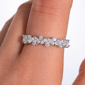 May include: A silver ring with a floral design, set with multiple clear, sparkling gemstones. The ring is worn on a finger, showcasing the intricate arrangement of the stones. The gemstones are a mix of round and marquise cuts.
