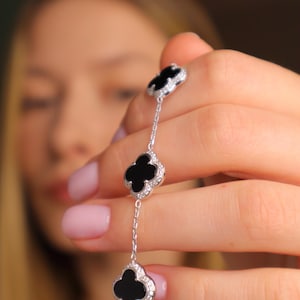 Puede incluir: Una pulsera de plata con tres colgantes negros en forma de trébol, cada uno bordeado con brillantes piedras blancas. La pulsera se sostiene, mostrando su delicado diseño y su elegante atractivo. El diseño de trébol es un motivo de joyería clásico.