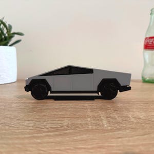 May include: A gray model truck with a sharp, angular, wedge-shaped body design, black wheels, and black window detailing. The vehicle features a flat roofline and a distinct geometric silhouette, presented in a side profile view on a flat surface.