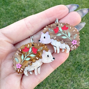 Può includere: Un paio di orecchini a forma di riccio marrone e bianco con fiori rossi e rosa e foglie verdi. Gli orecchini sono in acrilico e hanno una finitura lucida.