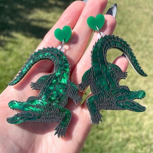 May include: A pair of green alligator earrings with a green heart charm. The alligator earrings are made of acrylic and have a detailed design.