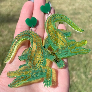 May include: A pair of green glitter alligator earrings with green heart charms. The alligator earrings are made of acrylic and have a shiny finish.